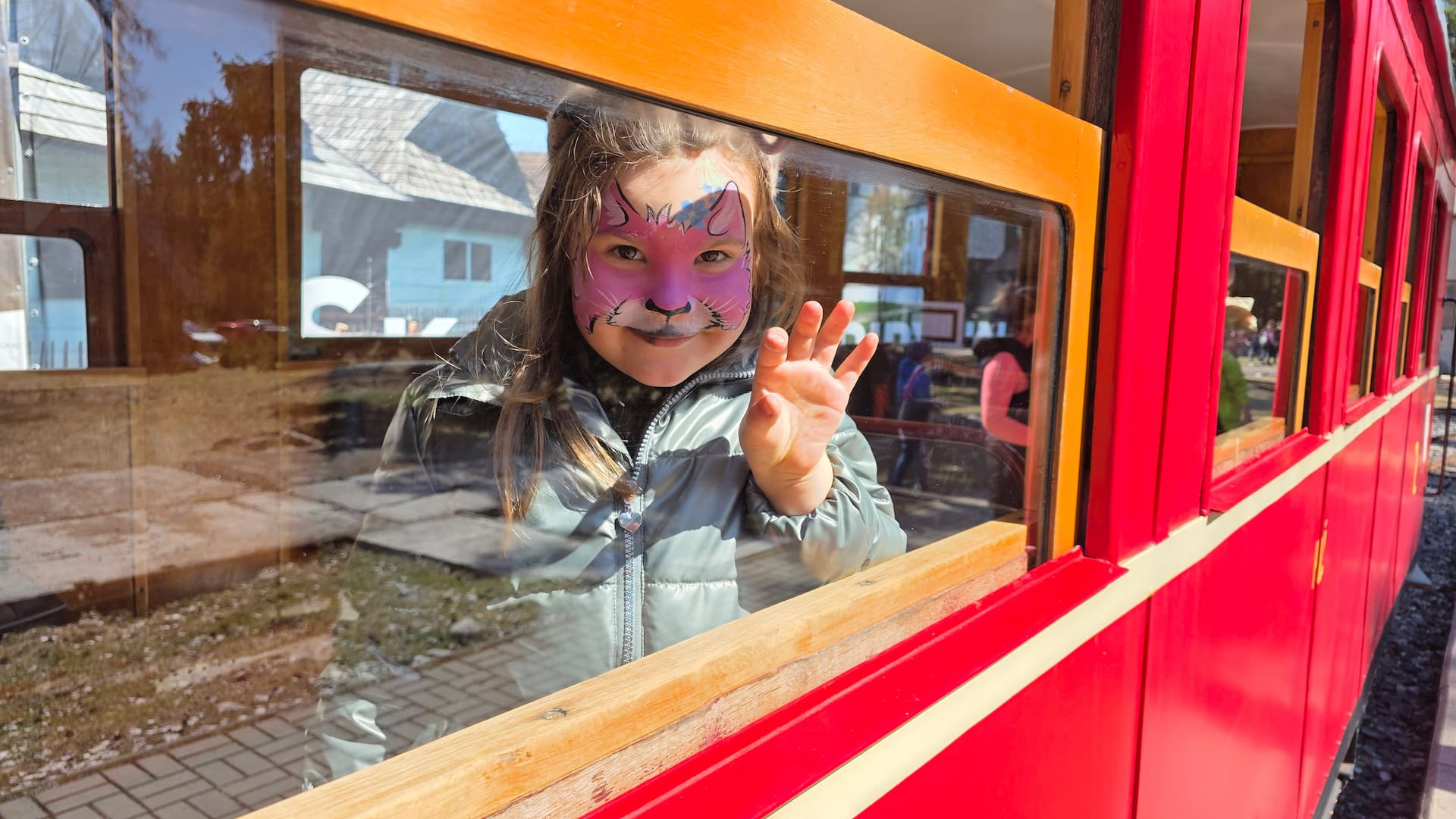 Dievčatko s maľovanou tvárou máva z okienka červeného vláčika v skanzene Pribylina
